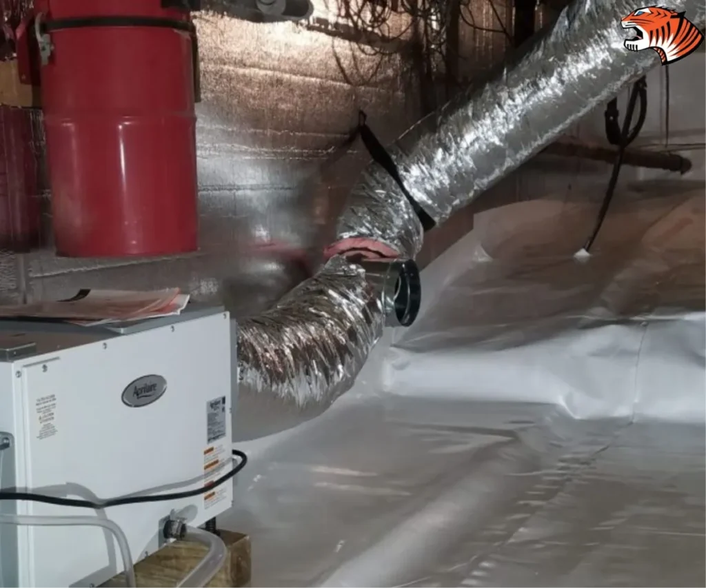 An Aprilaire dehumidifier unit connected to flexible metallic ductwork inside an encapsulated crawl space lined with white plastic vapor barrier.